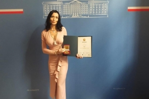 Agata Gacon w eleganckiej, różowej sukni trzyma dyplom i medal na tle ścianki z napisem „Śląski Urząd Wojewódzki w Katowicach”. Uhonorowana w oficjalnym wnętrzu, uśmiecha się z dumą, prezentując swoje wyróżnienie.