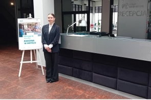 Ewa Raszka w eleganckim stroju hotelowym stoi przy recepcji Term Warmińskich. Obok znajduje się plakat promujący program "Rodzina w hotelu" organizowany przez SGH. Scena oddaje atmosferę profesjonalizmu i gościnności.
