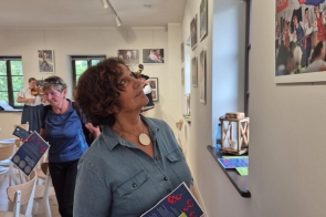 Janina Żagan ogląda z zainteresowaniem wystawę fotograficzną o folklorze. W dłoni trzyma program wydarzenia. W tle kobieta gra na skrzypcach, a inna uczestniczka przegląda materiały. Wnętrze jasne, pełne regionalnych akcentów.
