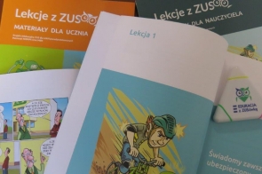 Rozsypane na biurku broszury „Lekcje z ZUS” — pomarańczowa dla ucznia i granatowa dla nauczyciela. Na wierzchu otwarta książeczka z napisem „Lekcja 1” i ilustracją chłopca jadącego na rowerze. Obok widać fragment komiksu oraz trójkątną, białą zakładkę z logo sowy „Edukacja z ZUSówką”.