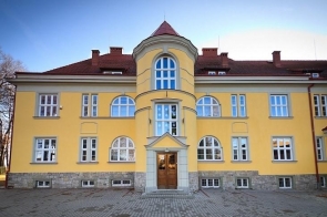 Symetryczny, dwukondygnacyjny Budynek główny Zespołu Szkół Przyrodniczo-Technicznych w Międzyświeciu, o żółtej elewacji z jasnymi obramowaniami okien, czerwonym dachem i centralną, lekko wysuniętą wieżą z półokrągłym zwieńczeniem; przed wejściem niewielki plac z kostki brukowej. 