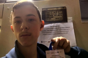 Chłopak robi zdjęcie typu selfie i pokazuje identyfikator z napisem „Staż uczniowski”. W tle widoczna jest maszyna oraz kartka informacyjna dotycząca projektu.