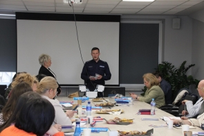 Policjant prowadzi spotkanie w sali konferencyjnej, mówiąc do grupy osób siedzących przy stole. Przed nim ekran projekcyjny, na stole materiały, przekąski i napoje. Uczestnicy słuchają, notują lub przeglądają broszury.