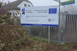 Metalowe ogrodzenie z zamontowaną tablicą informacyjną o dofinansowaniu z Unii Europejskiej. W tle budynek muzeum, krzewy i drzewa bez liści. Po prawej stronie widoczny fragment bramy i znak drogowy.