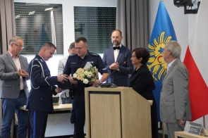 Podczas uroczystości w sali sesyjnej dwóch policjantów odbiera gratulacje i kwiaty z rąk przedstawicieli władz. Obok mównicy stoi Janina Żagan, a w tle widoczne są flagi: województwa śląskiego i Polski.