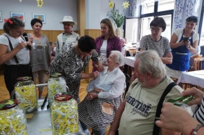 Grupa osób bierze udział w spotkaniu integracyjnym. Starsza kobieta siedzi, podczas gdy inna osoba przypina jej przypinkę. W tle inni uczestnicy obserwują i uśmiechają się. Na stole stoją słoiki wypełnione kolorowymi paskami papieru. 