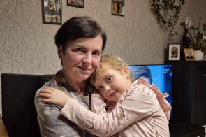 Starsza kobieta z krótkimi, ciemnymi włosami i łagodnym uśmiechem trzyma w ramionach małą dziewczynkę z jasnymi włosami. Dziewczynka, ubrana w różową bluzkę, przytula się z miłością do kobiety, spoglądając spokojnie przed siebie. W tle widać wnętrze mieszkania, na ścianie znajdują się zdjęcia oraz rośliny ozdobne. Atmosfera zdjęcia jest ciepła i rodzinna.
