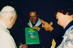 Janina Żagan wręcza papieżowi Janowi Pawłowi II książkę pt. „Ziemia Cieszyńska”. Obok stoją duchowny i kobieta z identyfikatorem. Wszyscy uśmiechnięci, skupieni na symbolicznym geście.