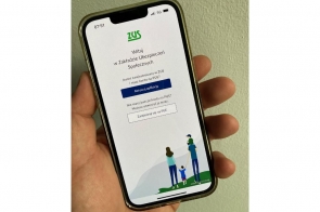Telefon trzymany w lewej dłoni wyświetla ekran powitalny aplikacji ZUS (Zakład Ubezpieczeń Społecznych). Na górze znajduje się logo ZUS i tekst powitalny. Poniżej widoczne są dwa przyciski: „Aktywuj aplikację” oraz „Zarejestruj się na PUE”. W dolnej części ekranu widać rysunek rodziny – dwoje dorosłych i dwójka dzieci.