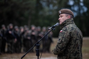 Żołnierz w mundurze polowym mówi do mikrofonu podczas zbiórki, w tle widoczna grupa wojskowych ustawionych w szeregu na otwartym terenie.