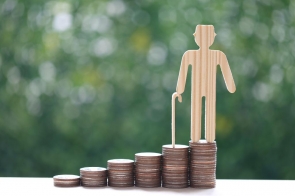 Drewniana figurka starszej osoby z laską stoi na najwyższym z kilku stosów monet ustawionych obok siebie w rosnącym układzie, na tle rozmytej zieleni.