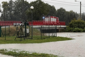 Straż pożarna stoi na moście podczas intensywnego deszczu. Woda z pobliskiego terenu zaczyna się rozlewać, co pokazuje zagrożenie powodziowe. W tle widać drzewa, a na pierwszym planie zielony teren ogrodzony siatką.