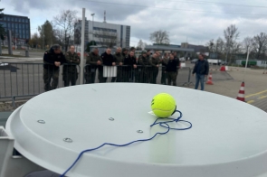 ChatGPT powiedział:  Żółta piłka tenisowa z przywiązanym niebieskim sznurkiem leży na białej antenie satelitarnej. W tle grupa umundurowanych osób stoi za barierką na placu, obok widoczne są pachołki drogowe i biurowce.