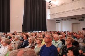 Sala pełna osób starszych, siedzących blisko siebie na krzesłach, uważnie patrzących w stronę sceny lub ekranu. Wnętrze jasne, z wysokim sufitem, dużymi zasłonami i balkonem u góry.
