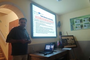 Osoba stoi po lewej przy łukowym przejściu. Na ścianie świeci ekran z prezentacją z ikonami i strzałkami. Przed nim biurko z laptopem i sprzętem; na blacie małe flagi. Na ścianie obok wisi mapa/obraz.