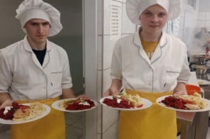Dwoje młodych kucharzy w białych fartuchach i czapkach kucharskich trzyma po dwa talerze z kolorowymi daniami. W tle widać kuchnię szkolną, uczniów przy pracy i parujący garnek na kuchence.