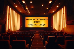 Duża, pusta sala kinowa z długimi rzędami foteli ustawionych amfiteatralnie, skierowanych w stronę szerokiego ekranu. Na ekranie wyświetlony jest napis „OTWARTY KONKURS OFERT ROZSTRZYGNIĘTY”. Wnętrze wypełnia ciepłe, pomarańczowe światło, które podkreśla spokojny i oficjalny charakter miejsca.
