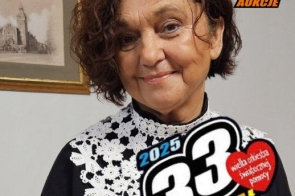 Janina Żagan, Starosta Cieszyński ubrana w elegancką czarną bluzkę z białymi, koronkowymi zdobieniami. W tle widoczna jest ramka z obrazem przedstawiającym budynek. W lewym dolnym rogu grafiki umieszczono logo 33. Finału Wielkiej Orkiestry Świątecznej Pomocy 2025. Atmosfera radosna i ciepła.