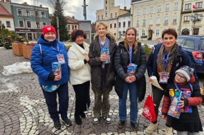 Grupa sześciu osób, w różnym wieku, druga od lewej – Janina Żagan, Starosta, stoi na brukowanym placu w centrum miasta. W rękach trzymają kolorowe puszki z logo Wielkiej Orkiestry Świątecznej Pomocy, uśmiechają się i mają przypięte identyfikatory. W tle widać kamienice, ratusz i dekoracje zimowe. Atmosfera pełna zaangażowania i życzliwości.
