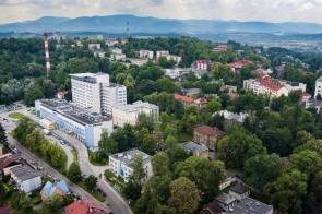 Widok z lotu ptaka na zabudowaną, zieloną okolicę Cieszyna z dużym kompleksem Szpitala Śląskiego w centrum kadru. W tle widać wzgórza, lasy, bloki mieszkalne i charakterystyczny komin w biało-czerwone pasy.
