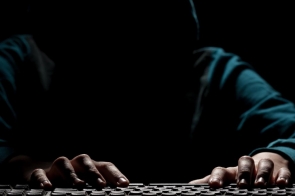 Osoba w kapturze siedzi przy komputerze w ciemnym otoczeniu, a oświetlenie podkreśla jedynie dłonie piszące na klawiaturze.