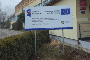 Poziomy baner informacyjny ustawiony na metalowych słupkach przed budynkiem użyteczności publicznej. Na banerze umieszczone są logotypy Funduszy Europejskich, Unii Europejskiej oraz Województwa Śląskiego. Widoczny jest napis „Dofinansowane przez Unię Europejską” oraz informacja o realizacji projektu. W tle znajduje się elewacja budynku z oknami, chodnik oraz teren zielony.