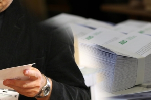 Osoba w ciemnym stroju trzyma w dłoni białą kartkę papieru, skupiając się na jej zawartości. W tle widoczny jest stos ułożonych równo dokumentów z zielonym logo, sugerujących oficjalny charakter. 