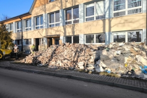 Przed dwukondygnacyjnym budynkiem z jasną elewacją i rzędami okien znajduje się dużo gruzu budowlanego z cegieł, fragmentów tynku, płyt oraz elementów sanitarnych. Przy wejściu do budynku stoją dwie osoby, jedna w odzieży roboczej. Po lewej stronie widoczna jest niewielka maszyna budowlana. Wzdłuż gruzu przebiega chodnik i jezdnia.