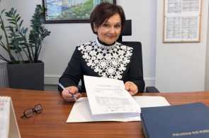 Janina Żagan Starosta Cieszyńska siedzi przy biurku w gabinecie i trzyma w dłoni dokument. Na biurku leżą segregatory, okulary oraz kalendarz, a w tle widać rośliny doniczkowe i obraz na ścianie.