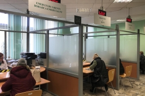 Wnętrze urzędu z boksami obsługi, gdzie klienci siedzą przy biurkach i załatwiają sprawy z pracownikami, w otoczeniu zielonych ścian, matowych przegród i elektronicznych wyświetlaczy numerków.