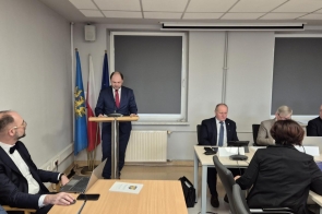 W Sali konferencyjnej Starostwa odbywa się XIX sesja Rady Powiatu Cieszyńskiego. Adam Śliż, dyrektor Szpitala Śląskiego w Cieszynie, ubrany w granatowy garnitur i czerwony krawat, stoi przy mównicy, przemawiając do obecnych. W tle widoczne są flagi narodowe oraz urzędowe. Przy stołach zasiadają radni w eleganckich strojach, skupieni na prezentowanych treściach. Atmosfera formalna i poważna.