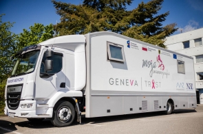 Biały mammobus — mobilna pracownia mammografii — zaparkowany na słonecznym parkingu. Na boku widoczne logotypy,  w tle zielone drzewa, fragment budynku i błękitne niebo.