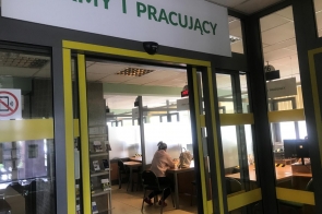 Przeszklone drzwi prowadzą do urzędowego pomieszczenia z oznaczeniem "firmy i pracujący", w środku przy biurku siedzi kobieta, odwrócona tyłem do obiektywu.