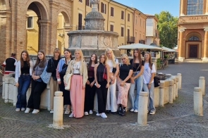 Grupa nastolatek stoi wokół kamiennej, zabytkowej fontanny na brukowanym placu. Wokół arkady z cegły, jasne kamienice, parasole kawiarniane i fasada kościoła.