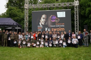 Grupa młodzieży oraz dorosłych stoi na trawie przed dużym ekranem z napisem „Śladami Zofii Kossak-Szatkowskiej. Ku swoim”. Wiele osób trzyma dyplomy. Obok znajduje się czarny namiot z napisem Lasów Państwowych. Atmosfera jest radosna i uroczysta.
