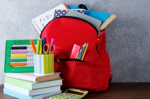 Czerwony plecak wypchany zeszytami, książkami i przyborami szkolnymi stoi na drewnianym stole obok stosu kolorowych książek, kubka z długopisami, liczydła i kalkulatora.