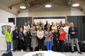 Grupowe zdjęcie uczestników spotkania, ustawionych na scenie w dwóch rzędach; większość uśmiechnięta, w centrum kobieta trzyma bukiet kwiatów. W tle ekran i zasłony sceniczne, po lewej roll-up z herbem Powiatu Cieszyńskiego. Nowoczesna sala z widoczną konstrukcją drewnianego stropu.