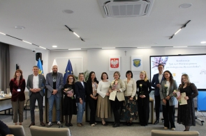 Grupa osób stojących w sali sesyjnej i pozujących do wspólnego zdjęcia. Część z nich trzyma kwiaty i dyplomy, a w tle widoczne są flagi, herby oraz ekran z prezentacją.