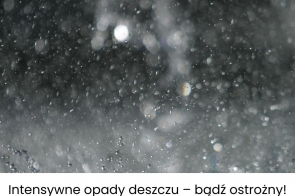 Rozmyte krople deszczu spadające w ciemności tworzą efekt mgiełki i refleksów świetlnych. U dołu białe tło z czarnym napisem: „Intensywne opady deszczu – bądź ostrożny!”.