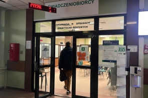 Wejście do sali obsługi ZUS z napisem „ŚWIADCZENIOBIORCY” i „Informacja w sprawach emerytalno-rentowych”. Mężczyzna w ciemnej kurtce wchodzi przez szklane drzwi, trzyma torbę. W środku widać krzesła, okienka, elektroniczny wyświetlacz kolejkowy oraz plakaty informacyjne na szybach.