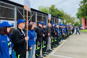 Grupa młodych osób w jednakowych strojach i niebieskich czapkach stoi w szeregu na zewnątrz, trzymając tabliczki z nazwami szkół. Uczestniczą w Olimpiadzie „Buduj z Pasją”.