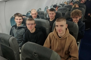 W rzędach szarych foteli samolotu siedzi sześciu nastolatków, większość w ciemnych bluzach; najbliżej po prawej chłopak w brązowej kangurce , obok uśmiechnięci koledzy, a za nimi kolejne rzędy pasażerów