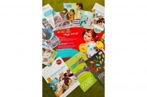 Kolorowe broszury edukacyjne rozłożone na zielonym tle przedstawiają materiały poświęcone emocjom, przemocy, asertywności, fonoholizmowi i rodzinie. Część z nich zawiera scenariusze zajęć, ilustracje dzieci i edukacyjne grafiki.