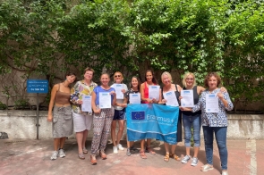Dziesięć uśmiechniętych kobiet pozuje na dworze z certyfikatami, a pośrodku trzymają niebieską flagę Erasmus+. Za grupą zielona ściana pnączy, słoneczna, radosna atmosfera.