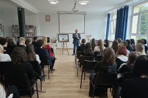 Na zdjęciu widać grupę uczniów siedzących w sali, którzy uczestniczą w spotkaniu z okazji Światowego Dnia Turystyki. Na środku, przed grupą, stoi prelegent w garniturze, który przemawia do zebranych. W tle widać ekran projekcyjny i tablicę informacyjną z napisem "Światowy Dzień Turystyki". Sala jest jasna, z dużymi oknami i regałami z książkami, co nadaje miejscu atmosferę nauki i skupienia.