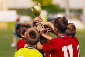 Grupa małych chłopców na boisku unosi razem złoty puchar. Czerwone koszulki piłkarskie, numery na plecach, medale na szyjach; jeden w żółtej bluzie. W tle rozmyta murawa i jasny dzień, euforia po wygranej. Dłonie na pucharze, miny skupione. W górze!