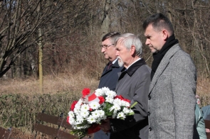 Trzech mężczyzn w eleganckich płaszczach i szalikach stoi w skupieniu – od prawej: Jacek Wiśniewski, sekretarz Powiatu, Stanisław Kubicius, Przewodniczący Rady Powiatu i Jerzy Pilch, członek Zarządu Powiatu. Stanisław Kubicius trzyma biało-czerwony wieniec z kwiatami, przewiązany wstążką w barwach narodowych. W tle ławki, przycięte krzewy i bezlistne drzewa, sugerujące chłodną porę roku.