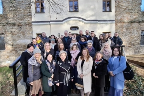 Grupa ludzi pozuje na tle Dworu Kossaków o jasnej fasadzie z widocznymi oknami i fragmentami ceglanej ściany. Wszyscy ubrani w jesienne płaszcze, uśmiechają się. Scena odbywa się w słoneczny dzień, w otoczeniu suchych liści i bezlistnych gałęzi. Atmosfera przyjazna i formalna.