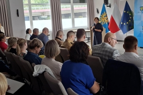 Pełna sala konferencyjna, uczestnicy siedzą w rzędach, zwróceni w stronę mównicy. Przy pulpicie przemawia Janina Żagan Starosta Cieszyński, w tle stoją flagi Polski, Unii Europejskiej i flaga regionalna oraz ścianka z herbem powiatu.