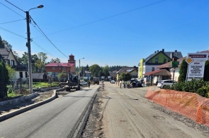 Zdjęcie przedstawia fragment ulicy Katowickiej w trakcie remontu w miejscowości Pogwizdów, gmina Hażlach. Widoczne są prace drogowe oraz maszyny budowlane na nawierzchni, a także wyznaczone strefy robót zabezpieczone pomarańczową siatką. Po obu stronach ulicy znajdują się budynki mieszkalne i usługowe, w tym restauracja „U Klimosza”. W tle widoczny jest również wiejski krajobraz i budynek strażnicy z czerwoną wieżyczką. 